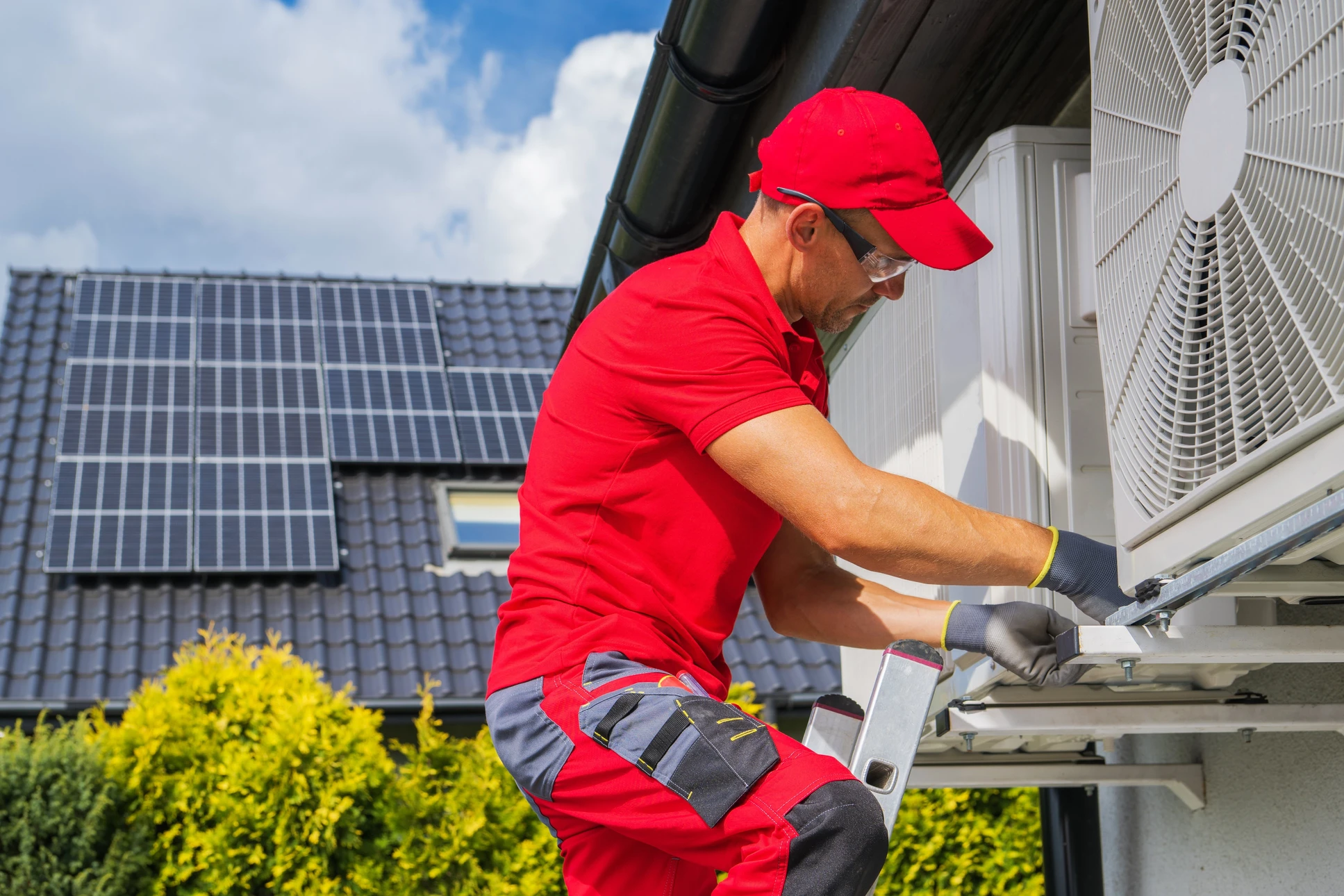 Technicien en uniforme rouge travaillant sur une unité de climatisation extérieure dans un environnement résidentiel avec panneaux solaires visibles sur toiture