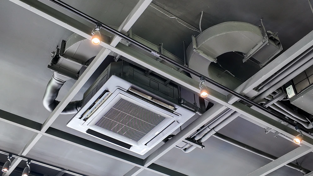 Plafond technique industriel avec système de climatisation cassette encastré, conduits de ventilation métalliques apparents et éclairage sur rails