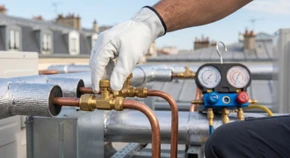 Photographie professionnelle d'une main gantée de blanc manipulant avec précision une valve de sécurité ou un raccord de tuyauterie en cuivre sur une installation frigorifique. L'image montre de près l'expertise technique, avec des outils métalliques spécialisés visibles en arrière-plan flou. L'éclairage naturel révèle la qualité et la propreté de l'installation, symbolisant la rigueur et la conformité technique garanties par la certification.